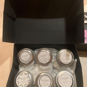 May include: A black box containing six silver-lidded tins of scented candles. Each tin has a white label with text, including "Actum Surge", "Healing Word", "Magic Intense", "Sneak Attack", and "Wild Shape". The candles are nestled in white tissue paper.