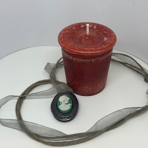 Op de afbeelding: Een rode kaars in een glazen pot met een witte lont. Een groene cameo-hanger met een wit silhouet van een vrouwenhoofd zit aan een bruin en grijs koord.