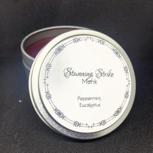 May include: A silver-colored round tin with the lid partially open, revealing a red interior. The lid features the text "Stunning Strike Monk" and the scent notes "Peppermint, Eucalyptus" in a decorative font.