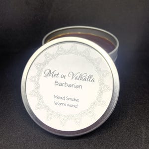 May include: A silver tin with a lid featuring the text "Met in Valhalla Barbarian". The lid also includes the words "Mead, Smoke, Warm wood" and an ornate border design. The tin is open, revealing a dark brown substance.