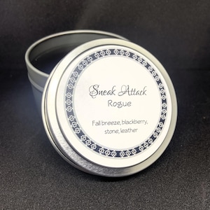 May include: A silver-colored tin with a lid featuring the text "Sneak Attack Rogue." The label also lists scent notes: fall breeze, blackberry, stone, and leather. The tin is open, revealing the dark interior.