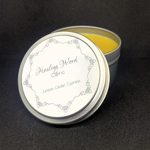 May include: Open silver tin with a yellow balm inside. The lid, slightly ajar, displays the text "Healing Word Cleric" within a decorative border, with "Juniper, Cedar, Cypress" below. The background is black.