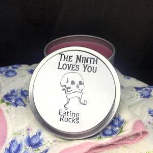 May include: A round, silver tin with a white lid featuring a skull illustration and the text "THE NINTH LOVES YOU" and "Eating Rocks" in black. The tin contains a dark pink substance and rests on a floral patterned cloth.