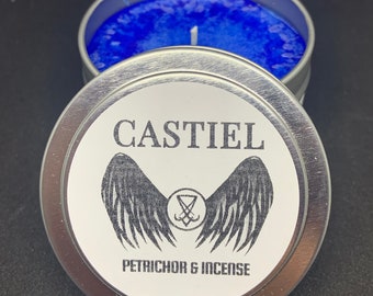 Op de afbeelding: Een zilveren blikje met een blauwe wasvulling. Het etiket op het blikje staat "CASTIEL" met een gestileerd engelenvleugelontwerp en de tekst "PETRICHOR & INCENSE".