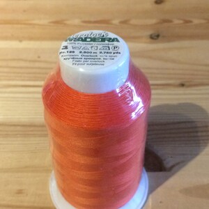 Könnte beinhalten: Orangefarbene Nähgarnrolle mit der Aufschrift "Madeira" und dem Text "100% Polyester / полиэфир No. 125 2.500 m 2.750 yds Kernzwim Overlock cure spun кручёные армиров, нитки Filato per overlock Fil pour surjeteuse"