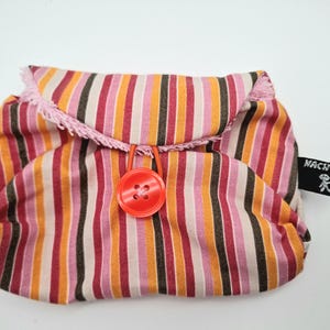 Könnte beinhalten: Eine gestreifte Stofftasche mit vertikalen Streifen in Rosa-, Orange- und Brauntönen. Die Tasche hat eine rosa Fransenborte und einen großen roten Knopfverschluss. Ein kleines schwarzes Etikett mit weißem Text und einer Strichmännchenfigur ist angebracht.