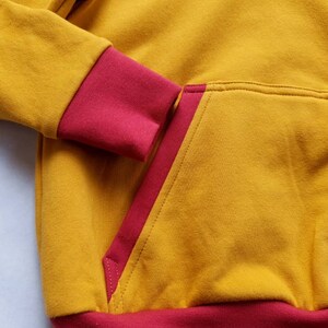 May include: A mustard yellow hoodie with a red trim around the hood, cuffs, and pocket. The hoodie has a kangaroo pocket with a red trim.