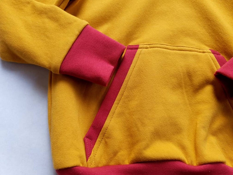 May include: A mustard yellow hoodie with a red trim around the hood, cuffs, and pocket. The hoodie has a kangaroo pocket with a red trim.