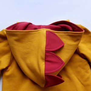 May include: A mustard yellow hoodie with a red dinosaur-shaped hood. The hood has a series of red spikes on the back.