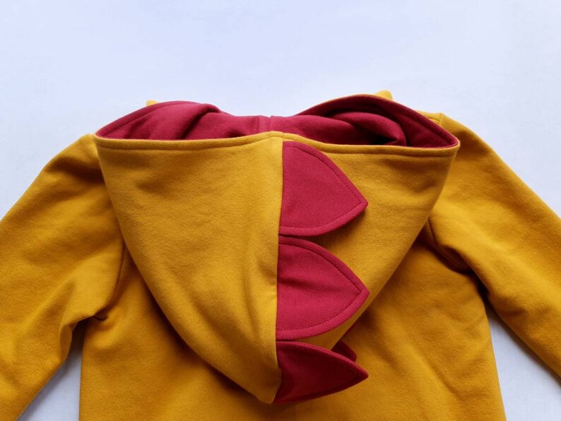 May include: A mustard yellow hoodie with a red dinosaur-shaped hood. The hood has a series of red spikes on the back.