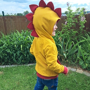 May include: A child wearing a yellow hooded sweatshirt with a red dinosaur-shaped hood. The child is standing in a grassy area with a wooden fence in the background.