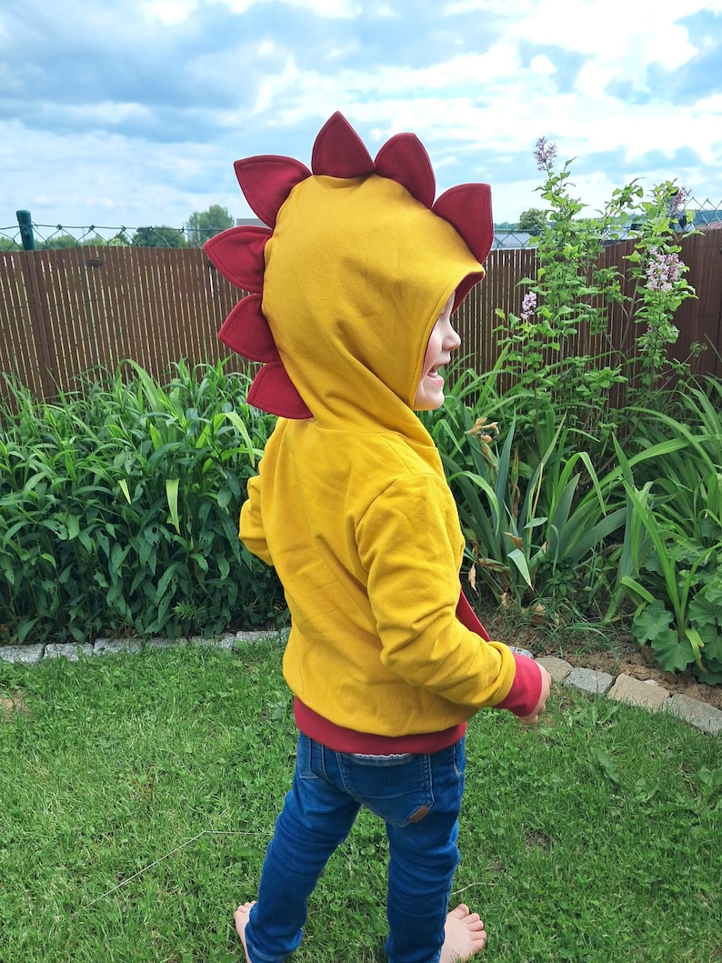 May include: A child wearing a yellow hooded sweatshirt with a red dinosaur-shaped hood. The child is standing in a grassy area with a wooden fence in the background.
