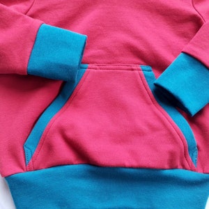 May include: A red hooded sweatshirt with teal blue trim around the cuffs and bottom hem. The sweatshirt has a single pocket on the front.
