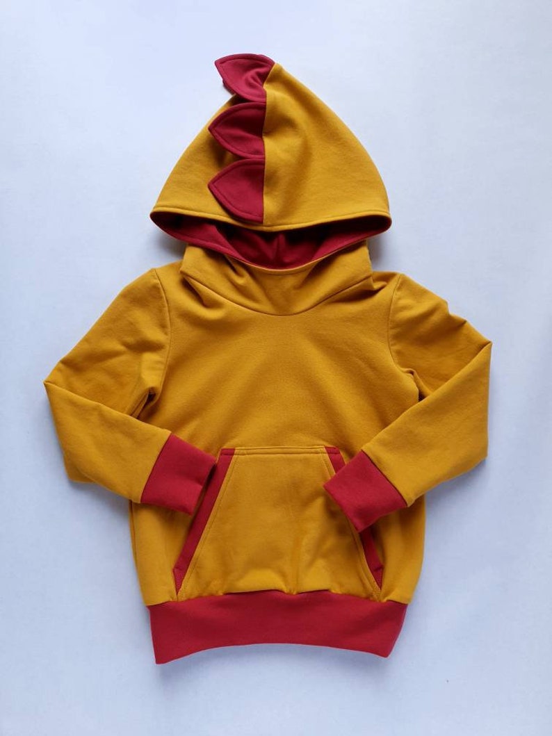 May include: A yellow hoodie with a red dinosaur-shaped hood and red trim on the sleeves and bottom. The hoodie has a kangaroo pocket.