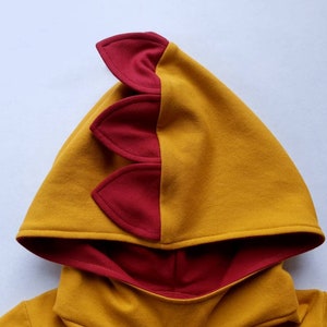 May include: A yellow hooded sweatshirt with a red dinosaur-shaped hood. The hood has three red spikes on the back.