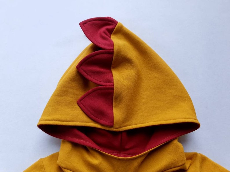 May include: A yellow hooded sweatshirt with a red dinosaur-shaped hood. The hood has three red spikes on the back.