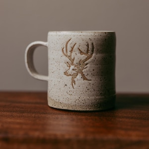 May include: A speckled ceramic mug with a deer head design. The mug is off-white with a brown deer head etched into the side. The mug has a handle and is sitting on a wooden surface.