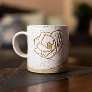 May include: A white ceramic mug with a floral design. The mug has a speckled finish and a tan-colored base. The flower is embossed in a light brown color. The mug has a curved handle.