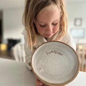 以下が含まれることがあります： 茶色の縁と「Juniper」という名前が茶色の文字で書かれた、斑点のある白いセラミックプレート。 プレートは人が持っています。