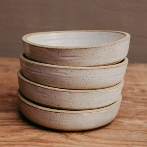 May include: A stack of four round ceramic bowls with a speckled off-white glaze. Each bowl features a slightly uneven, hand-formed rim and a textured surface with visible dark brown flecks throughout the light-colored clay body.
