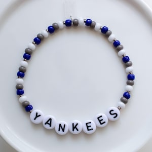 Op de afbeelding: Een witte kralenketting met grijze en blauwe kralen, die het woord "YANKEES" spelt in witte letters met zwarte letters.