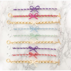May include: A set of nine bracelets with different colored chain links and bows. The bows are made of leather and come in a variety of colors, including purple, pink, blue, green, and white.