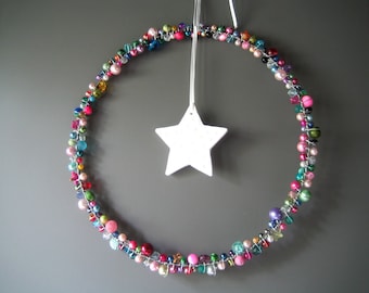 May include: A colorful beaded wreath with a white star-shaped ornament hanging in the center. The wreath is made of various sizes and colors of beads, including pink, blue, green, and yellow. The star is white and has a textured surface.