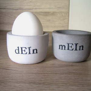 Könnte beinhalten: Zwei kleine, runde Eierbecher aus Beton. Einer ist weiß mit dem Wort "dEIn" in Schwarz bedruckt und hält ein weißes Ei. Der andere ist grau mit "mEIn" in Schwarz bedruckt.