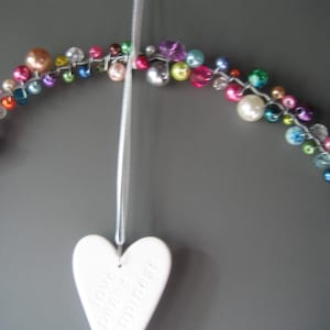 May include: A circular wire wreath adorned with a variety of multicolored beads in shades of pink, blue, green, purple, and white. A white ceramic heart hangs from a thin ribbon, featuring the embossed words love, peace, and happiness.