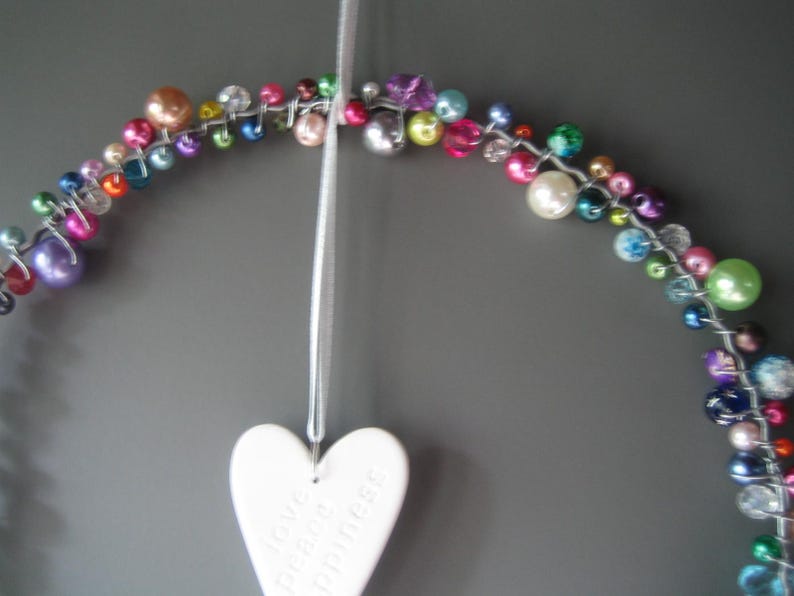 May include: A circular wire wreath adorned with a variety of multicolored beads in shades of pink, blue, green, purple, and white. A white ceramic heart hangs from a thin ribbon, featuring the embossed words love, peace, and happiness.