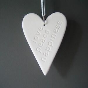 May include: A white heart-shaped ornament made of a smooth material, featuring the embossed words love, peace, and happiness stacked vertically. The heart hangs from a thin white ribbon attached to a silver metal jump ring at the top.