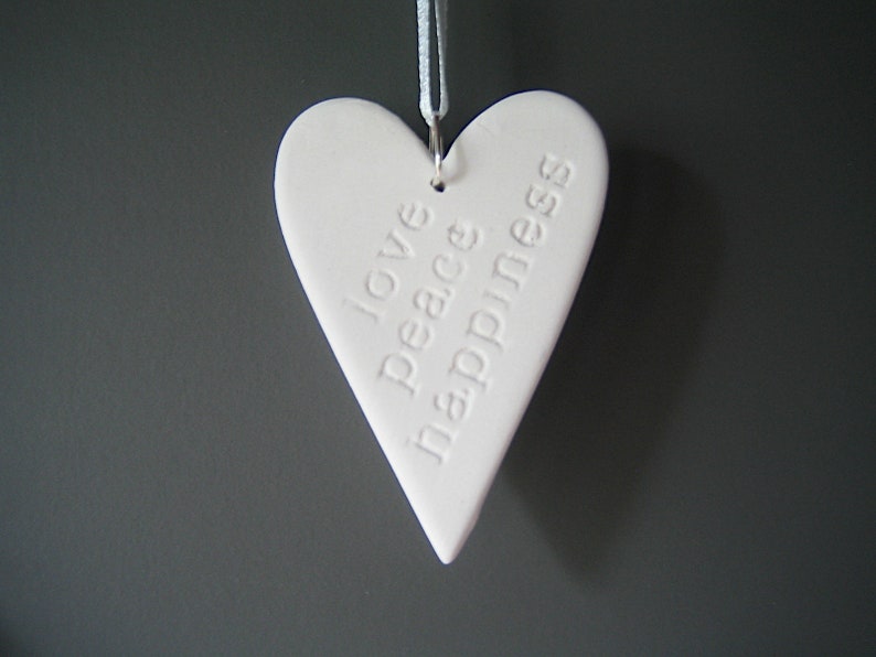 May include: A white heart-shaped ornament made of a smooth material, featuring the embossed words love, peace, and happiness stacked vertically. The heart hangs from a thin white ribbon attached to a silver metal jump ring at the top.