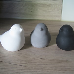 Könnte beinhalten: Drei Keramik-Vogelfiguren in Weiß, Grau und Schwarz. Die Vögel haben ein einfaches, abgerundetes Design. Die Figuren stehen auf einer Holzoberfläche.