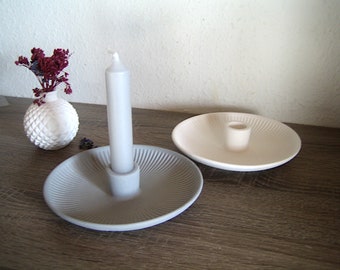 May include: Two ceramic candle holders with a ridged design. One is light gray with a white candle, and the other is white. A small white vase with a textured pattern holds dried flowers.