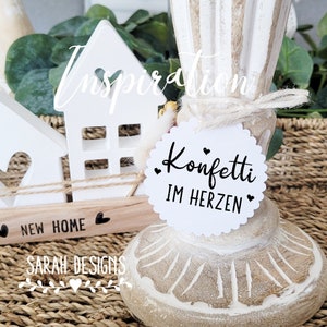 Könnte beinhalten: Weiße Holzhausdekoration mit dem Text "New Home" und einem kleinen Herz. Ein weißer runder Anhänger mit dem Text "Konfetti im Herzen" und zwei kleinen Herzen ist an einem weißen Holzkerzenhalter befestigt.