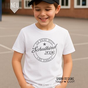 May include: A white t-shirt featuring a grey circular design with the text "ICH ROCKE JETZT Schulkind 2026 DIE SCHULE". The shirt is worn by a child wearing a black baseball cap. The text "SARAH DESIGNS" is at the bottom right.
