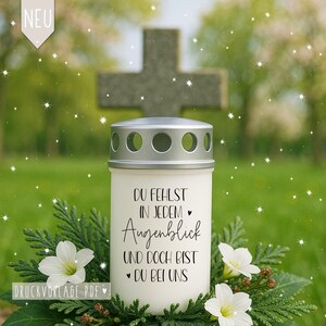 Op de afbeelding: Een witte herdenkingskaars met een zilveren deksel, met de tekst "DU FEHLST IN JEDEM Augenblick UND DOCH BIST DU BEI UNS". De kaars is omgeven door groen en witte bloemen, met een kruis op de achtergrond.