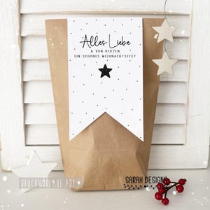 Könnte beinhalten: Eine braune Papiertüte mit einem weißen Banner mit dem Text "Alles Liebe & von Herzen EIN SCHONES WEIHNACHTSFEST" und einem schwarzen Stern. Zwei weiße Holzstern-Ornamente hängen über der Tüte. Die Worte "SARAH DESIGNS" sind sichtbar.