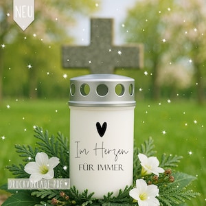 Op de afbeelding: Een witte herdenkingskaars met een zilveren deksel, met een zwart hart en de woorden "Im Herzen FÜR IMMER". De kaars is omgeven door groen en witte bloemen, met een kruis op de achtergrond.