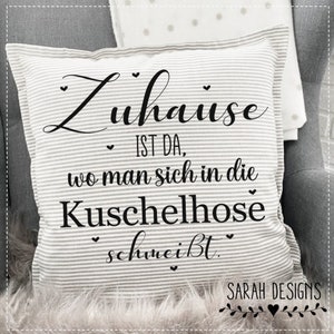 Könnte beinhalten: Ein weiß-graues, gestreiftes Kissen mit schwarzem Text, der "Zuhause ist da, wo man sich in die Kuschelhose schmeiBt." lautet. Der Text ist in einer kursivschriftlichen Schriftart und enthält kleine Herzen. Am unteren Rand des Kissens steht der Text "SARAH DESIGNS" mit einem kleinen Herzen.