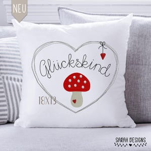 Op de afbeelding: Een wit kussen met een grijs hartontwerp en de tekst "Glückkind" in zwart. In het hart bevindt zich een rode en witte paddenstoel met een beige steel. Het kussen is 18x13 inch.