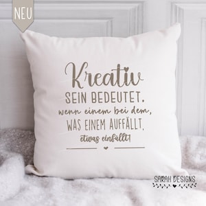 Puede incluir: Almohada blanca con texto marrón que dice "Kreativ Sein Bedeutet. Wenn einem bei dem, Was einem auffällt, etwas einfällt!" y un pequeño corazón en la parte inferior. La almohada tiene un borde marrón y el texto "SARAH DESIGNS" con un pequeño corazón en la parte inferior.