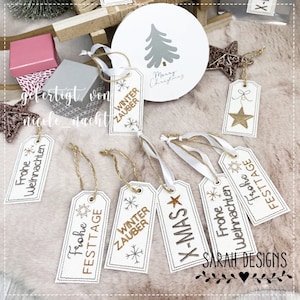 Peut inclure: Un ensemble de huit étiquettes cadeaux blanches avec des lettres dorées et des flocons de neige. Les étiquettes présentent diverses phrases sur le thème de Noël et de l'hiver, notamment "Frohe Weihnachten", "Winterzauber" et "X-MAS". Les étiquettes sont attachées avec de la ficelle et un ruban blanc.