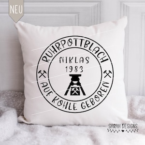 Puede incluir: Cojín cuadrado blanco con un diseño circular negro. El diseño incluye el texto "RUHRPOTTBLACH", "NIKLAS 1983" y "AUF KOHLE GEBOREN" alrededor de un gráfico de mina de carbón. El cojín tiene una etiqueta "NEU".