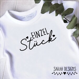 Puede incluir: Un body blanco para bebé con texto negro que dice "EINZEL STÜCK" con dos pequeños corazones a cada lado del texto. El texto está en una fuente cursiva. El body tiene una etiqueta que dice "Sarah Designs".