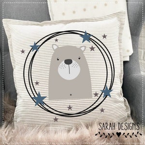 May include: A beige and white striped pillow with a brown bear face inside a circle of stars. The circle is outlined in black and has a blue star at the top. The text "SARAH DESIGNS" is at the bottom of the pillow.