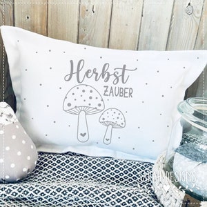 Op de afbeelding: Wit kussen met grijze tekst die "Herbst Zauber" zegt en twee grijze paddenstoel illustraties. Het kussen ligt op een grijs en wit gepatroneerd oppervlak.