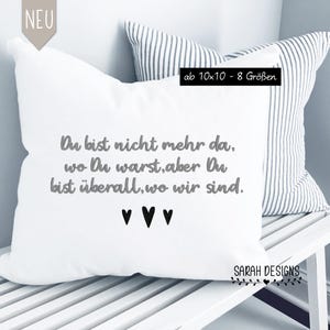 May include: White square pillow with gray text that reads "Du bist nicht mehr da, wo Du warst, aber Du bist überall, wo wir sind." Below the text are three black hearts. A second pillow with vertical stripes is behind it.