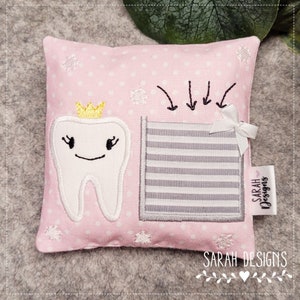 Puede incluir: Almohada rosa con un diente blanco bordado en ella que lleva una corona dorada. El diente sonríe y tiene pestañas. Hay un bolsillo a rayas gris y blanco en la almohada con flechas que apuntan hacia él. La almohada tiene una etiqueta blanca con el texto "SARAH DESIGNS" en ella.