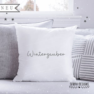 May include: White throw pillow with the text "Winterzauber" in gray script on a gray couch with striped pillows.